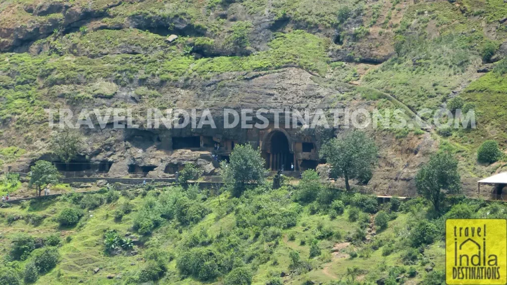 A view of Bhaja Caves in Lonavala with greenery, hillside trail, and rocky cliff face.